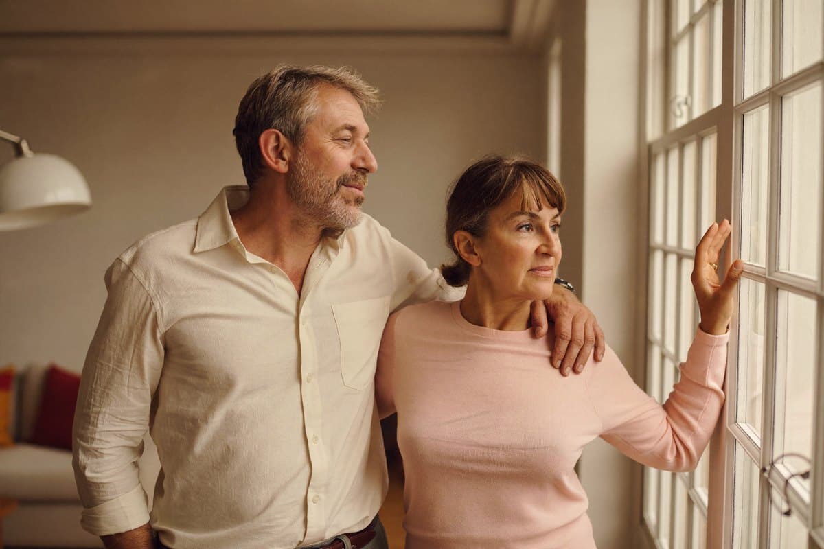 Couple in their fifties standing calmly at a window, looking out relaxed